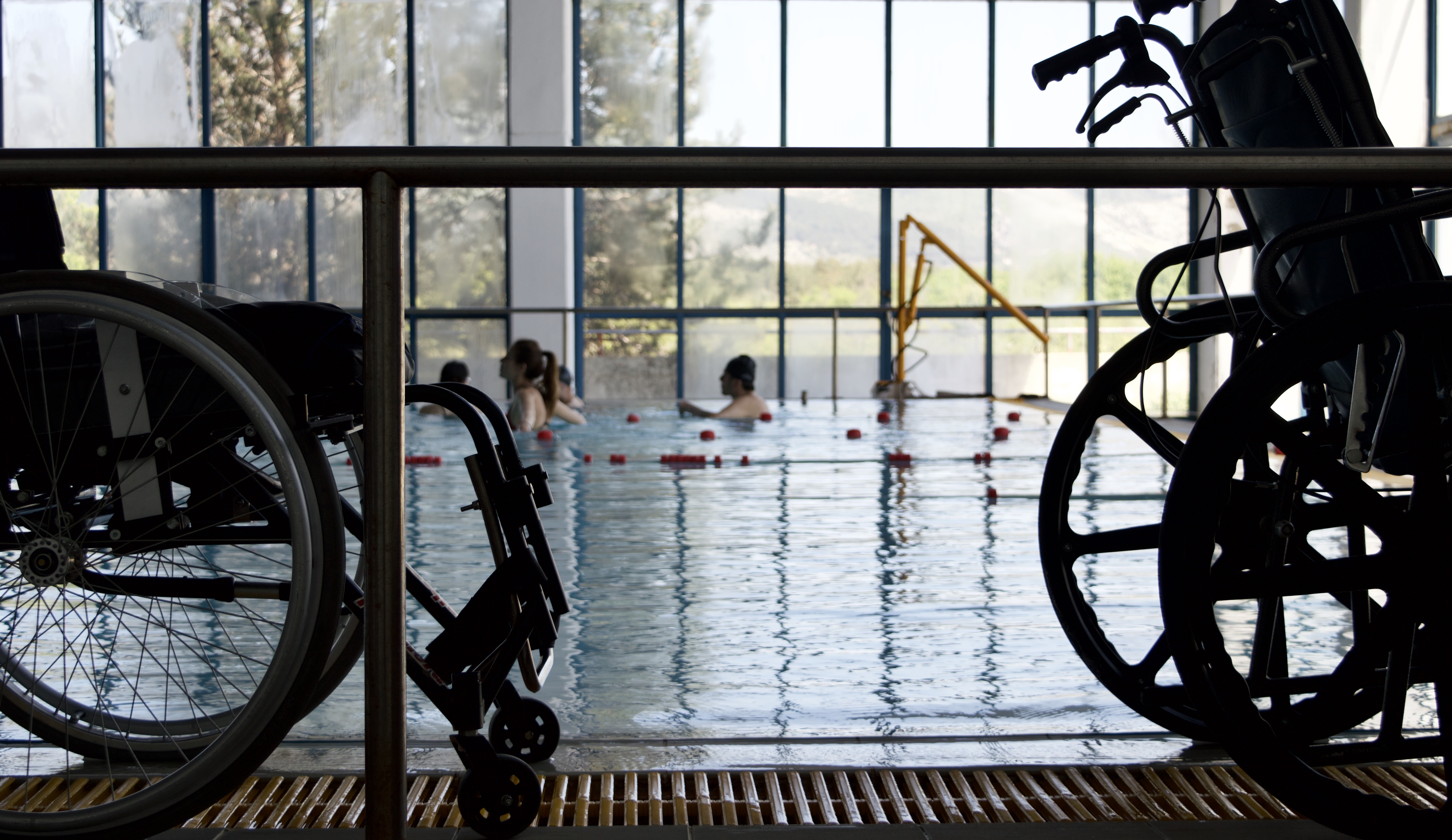 Cover. A couple of vacant wheelchairs next to an indoors swimming pool with people in it.